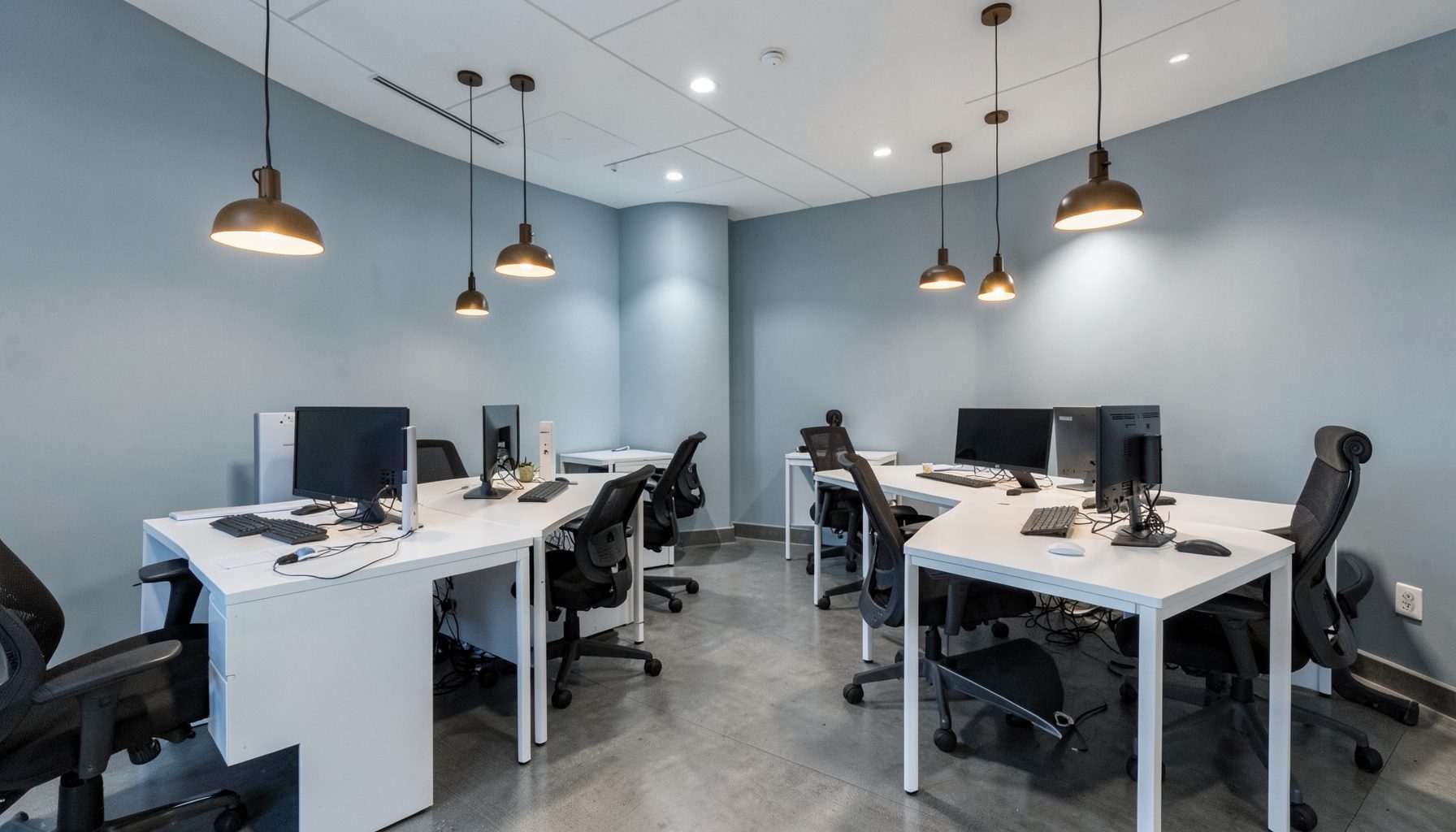 A sleek modern office interior with soft blue-gray walls, warm pendant lighting, and clean white desks arranged for collaborative work